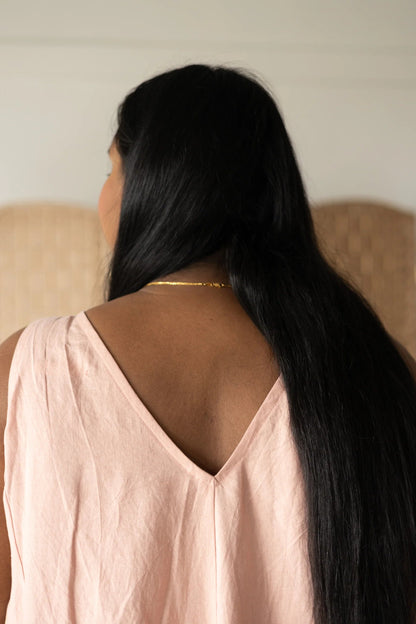 Back close up view of a woman wearing the Caroline Dress by Lights of All in Ballet Pink, a sleeveless dress linen maxi dress with a V-neck at the back. She is standing against a white wall with a woven screen behind her. 