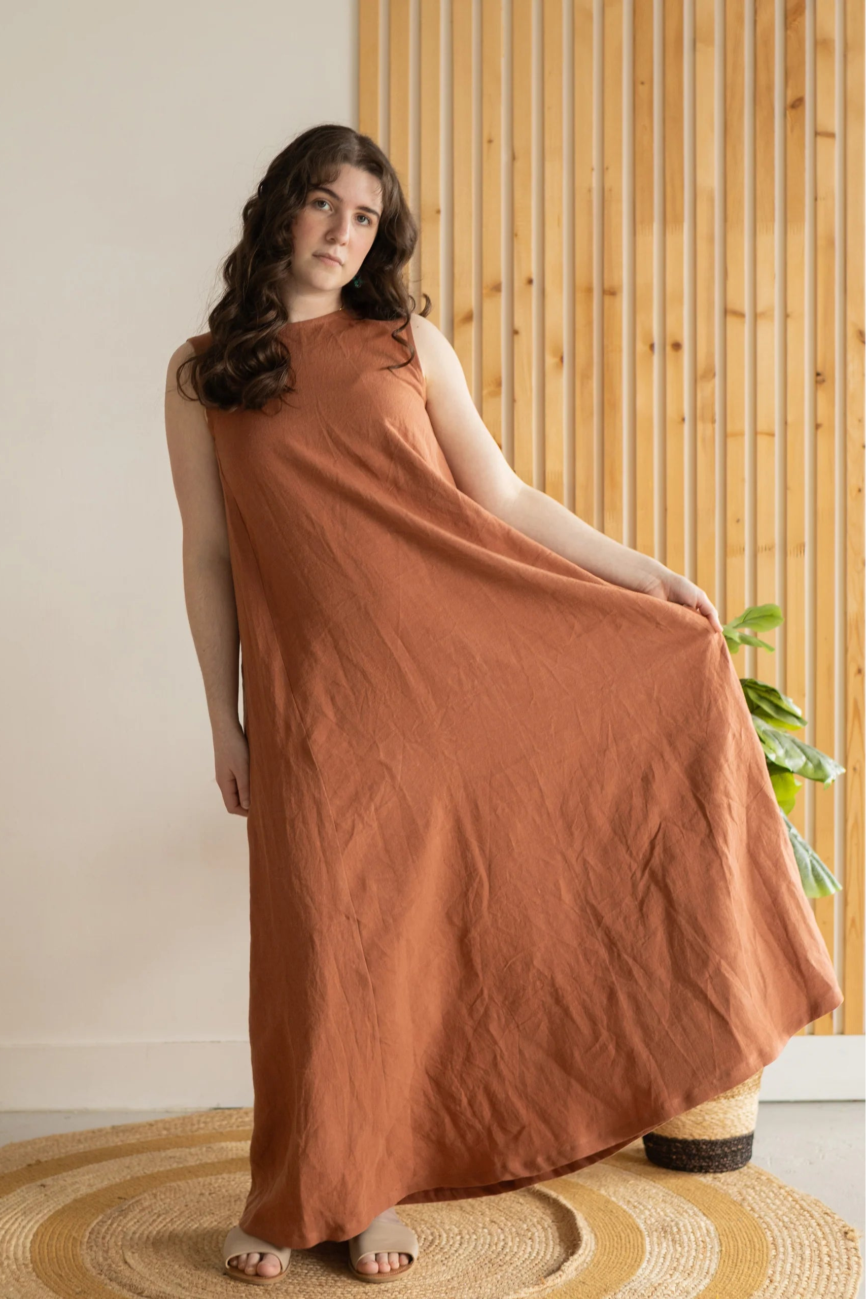 A woman wearing the Caroline Dress by Lights of All in Sedona, a sleeveless dress linen maxi dress with a V-neck at the back. She is standing against a white and wood panelled wall. 