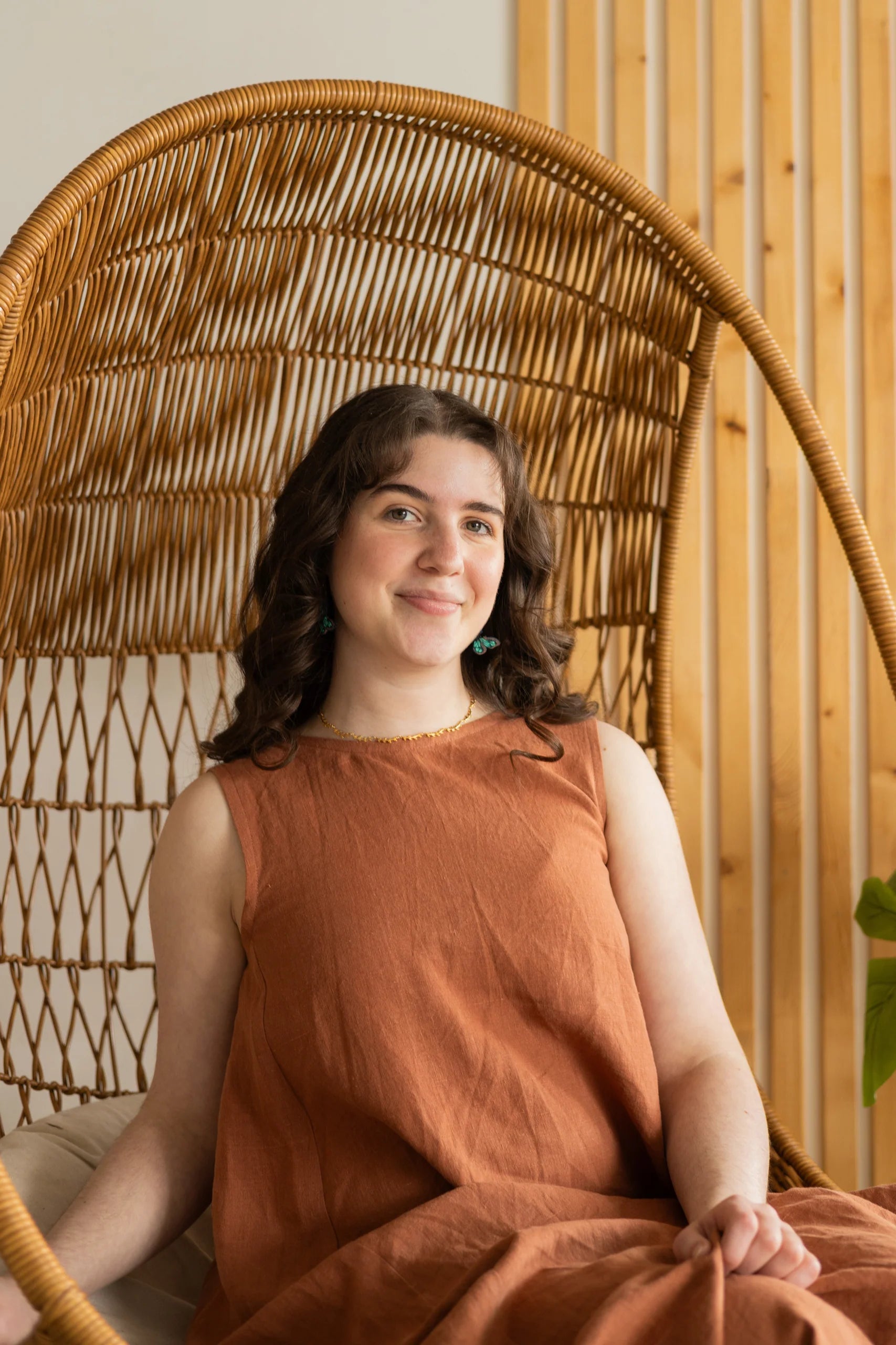 A woman wearing the Caroline Dress by Lights of All in Sedona, a sleeveless dress linen maxi dress with a V-neck at the back. She is seated in a woven chair. 