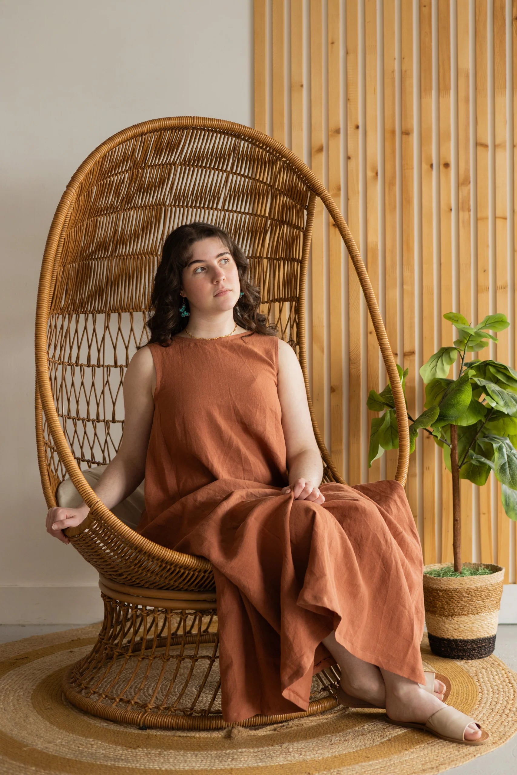 A woman wearing the Caroline Dress by Lights of All in Sedona, a sleeveless dress linen maxi dress with a V-neck at the back. She is seated in a woven chair
