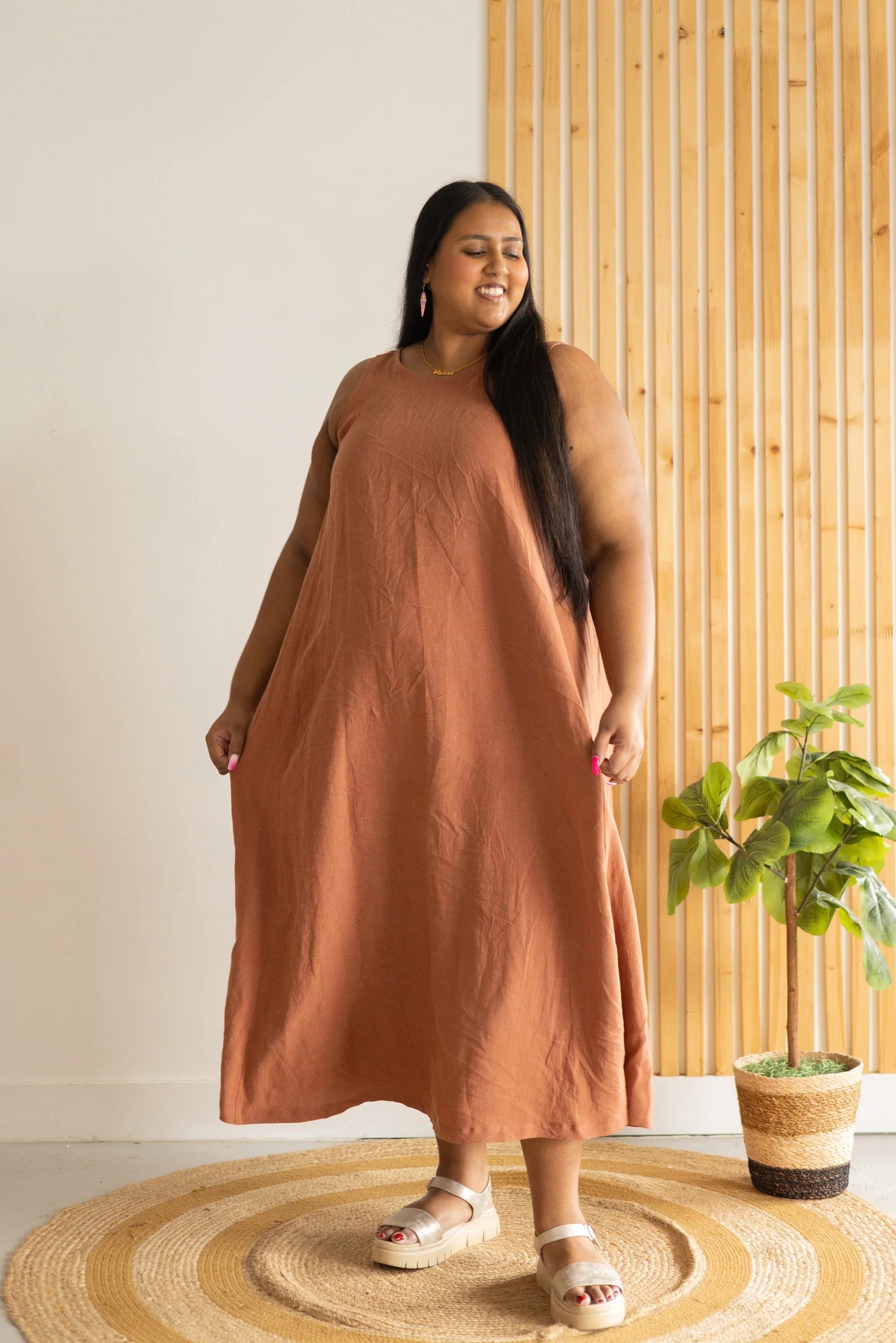 A woman wearing the Caroline Dress by Lights of All in Sedona, a sleeveless dress linen maxi dress with a V-neck at the back. She is standing against a white and wood panelled wall. 