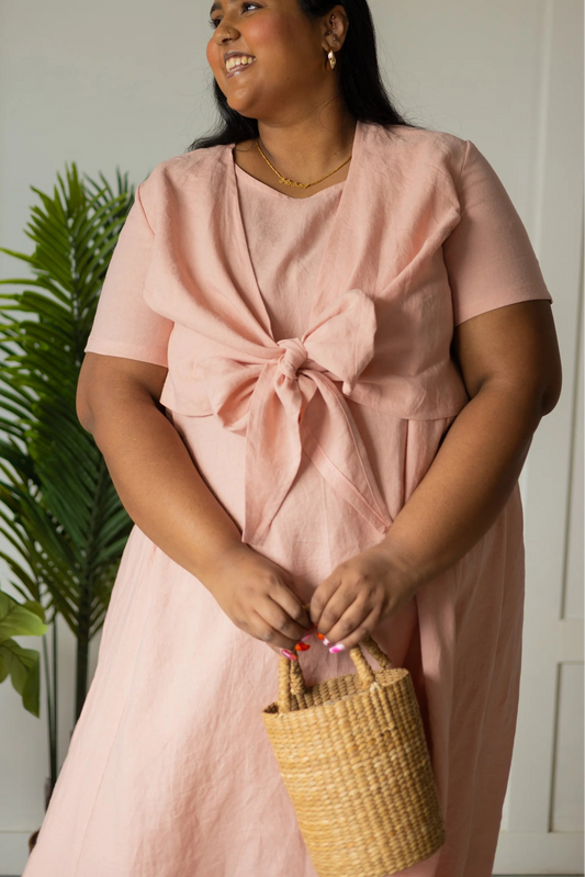 A woman wearing the Stella Top by Lights of All in Ballet Pink, a short sleeved linen wrap top that can be twisted or tied back or front. She is wearing it over the matching Caroline dress and standing in front of a plant holding holding a straw bag. 