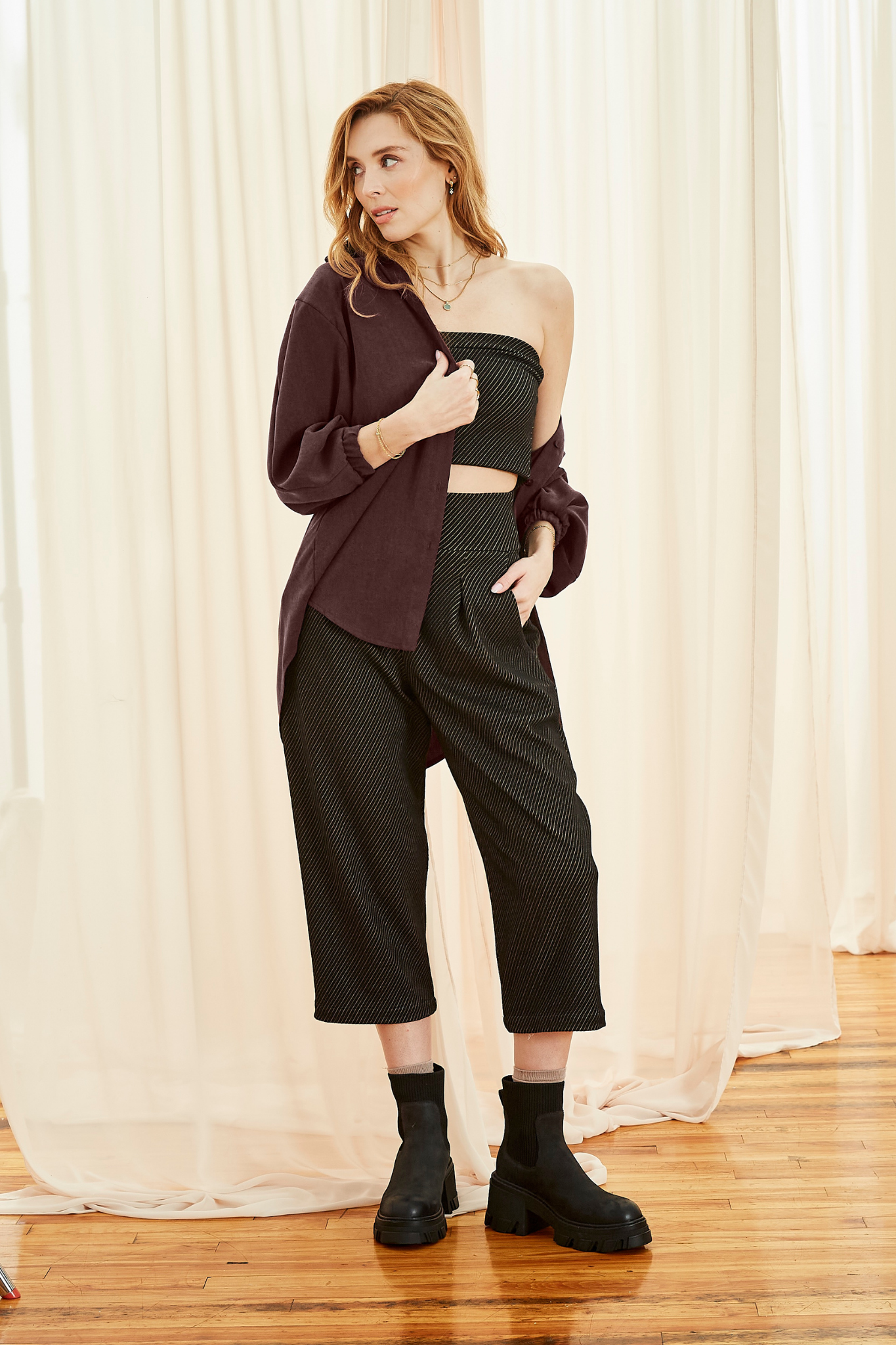 A woman wearing the Lucas Pants by Melow in Black, featuring a diagonal stripe texture, a high elastic waist, front pleats, and 3/4 length legs. She is wearing them with a tube top and ajn open burgundy shirt and and standing in front of a white curtain. 