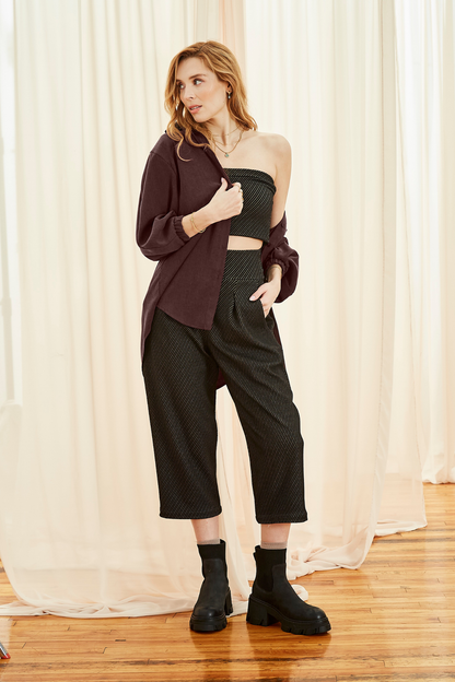 A woman wearing the Lucas Pants by Melow in Black, featuring a diagonal stripe texture, a high elastic waist, front pleats, and 3/4 length legs. She is wearing them with a tube top and ajn open burgundy shirt and and standing in front of a white curtain. 