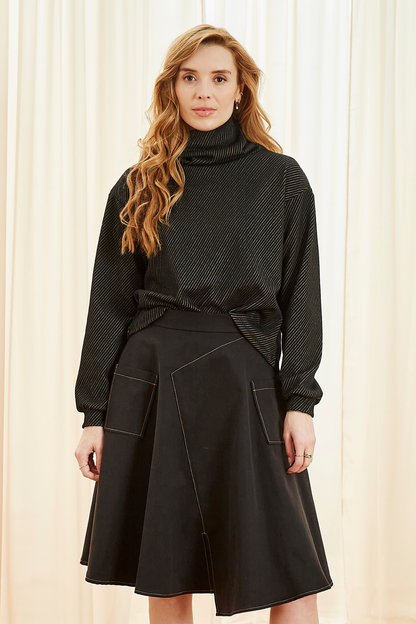 A woman wearing the Ludovic Sweater by Melow in Black, a square shaped turtleneck sweater with puffed sleeves that taper at the wrists and a diagonal stripe texture. She is wearing it with a skirt and standing in front of a curtain. 
