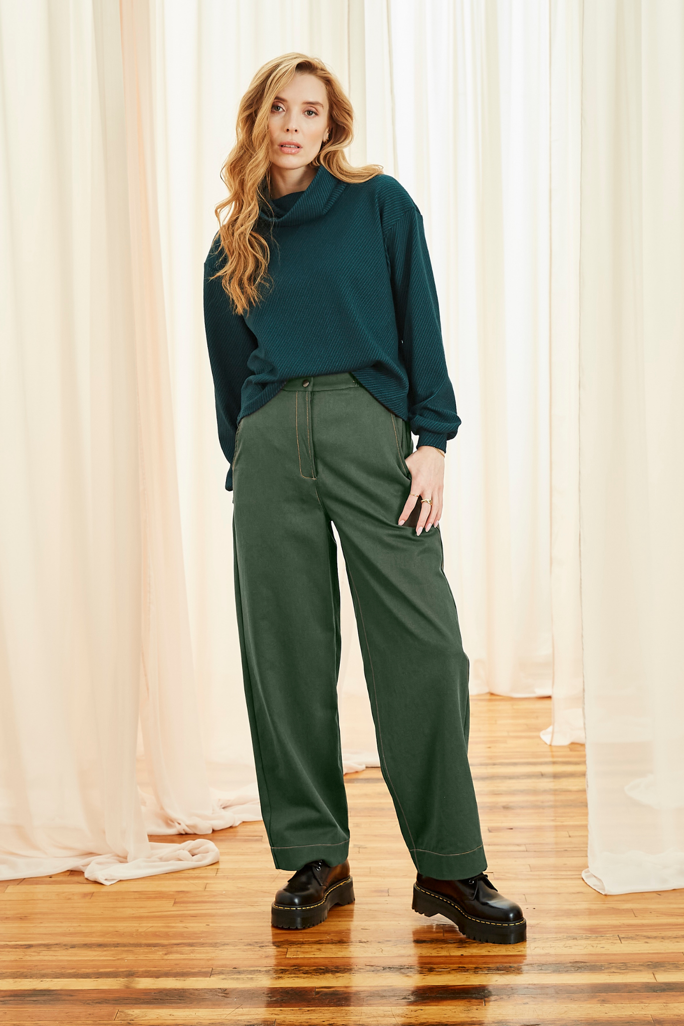 A woman wearing the Ludovic Sweater by Melow in Oil, a teal coloured square shaped turtleneck sweater with puffed sleeves that taper at the wrists and a diagonal stripe texture. She is wearing it with green pants and standing in front of a curtain. 
