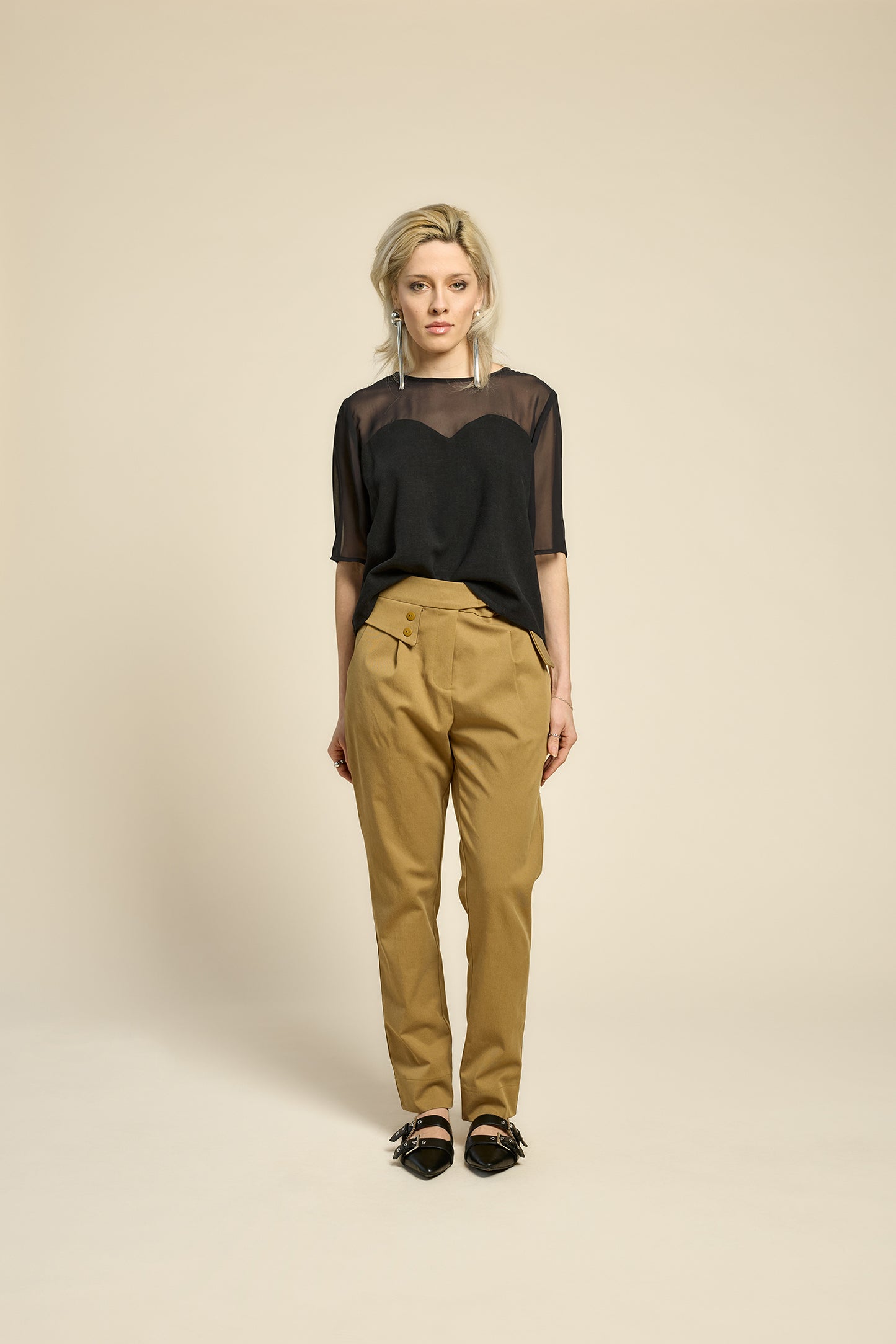 A woman wearing the L'Amour Top by Cokluch in Black, with a heart-shaped bodice with a sheer overlay and short sleeves. She is wearing it with beige pants against a beige background