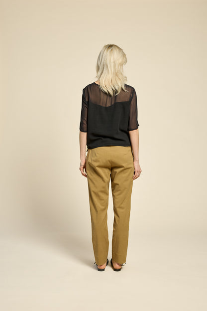 Back view of a woman wearing the L'Amour Top by Cokluch in Black, with a heart-shaped bodice with a sheer overlay and short sleeves. She is wearing it with beige pants against a beige background