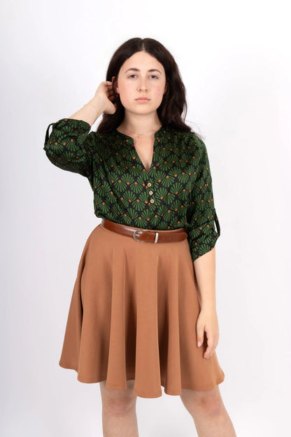 A women wearing the Leonie Blouse by Infime in Black with Green print, featuring a notched V-neckline, three button placket, and 3/4 sleeves. She is wearing it with a tan skirt and standing in front of a white background. 