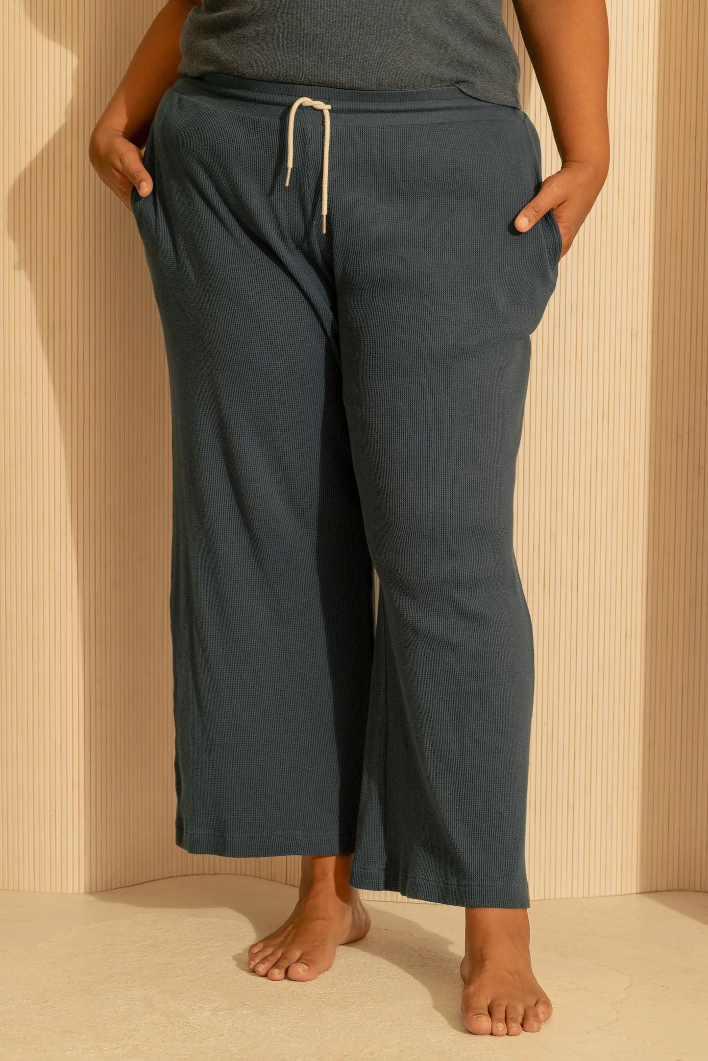 Waist down view of a woman wearing the Organic Cotton Lounge Pants from Lights of All in Deep Sea, waffle knit pants with an elastic and drawstring waist, straight legs, and pockets. She is wearing them with a charcoal tank top and standing against a cream wall. 