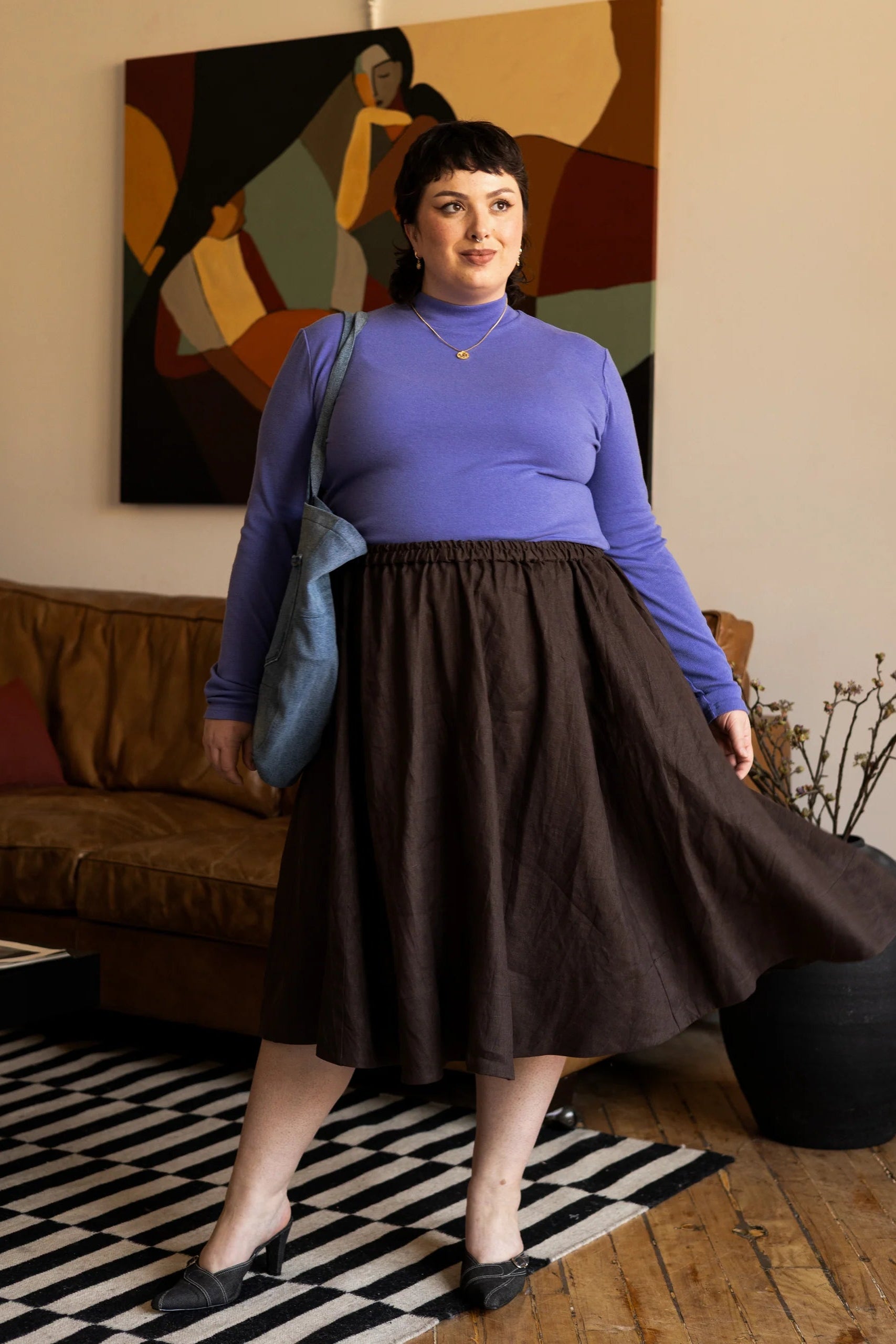 Woman wearing the Tuesday Skirt by Lights of All in Cocoa with the Essential turtleneck in Violet standing in a living room with abstract art on the wall.