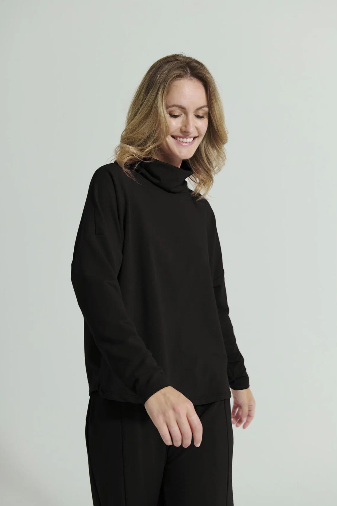 3/4 view of a woman wearing the Romaine Sweater by Message Factory in Black, a bamboo/organic cotton sweatshirt with a funnel neck , a loose fit, and long drop sleeves. She is wearing the matching Tadoussac Pants and standing in front of a white background. 