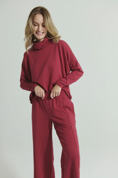 A woman wearing the Romaine Sweater by Message Factory in Raspberry, a bamboo/organic cotton sweatshirt with a funnel neck, a loose fit, and long drop sleeves. She is wearing the matching Tadoussac Pants and standing in front of a white background. 