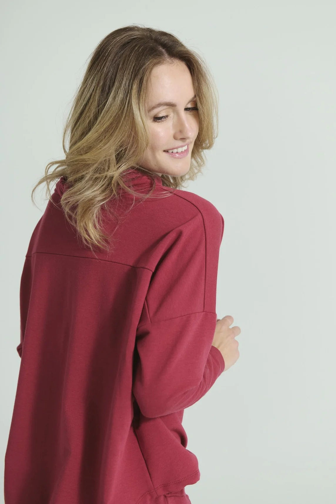 Back Waist up view of a woman wearing the Romaine Sweater by Message Factory in Raspberry, a bamboo/organic cotton sweatshirt with a funnel neck , a loose fit, and long drop sleeves. She is standing in front of a white background. 