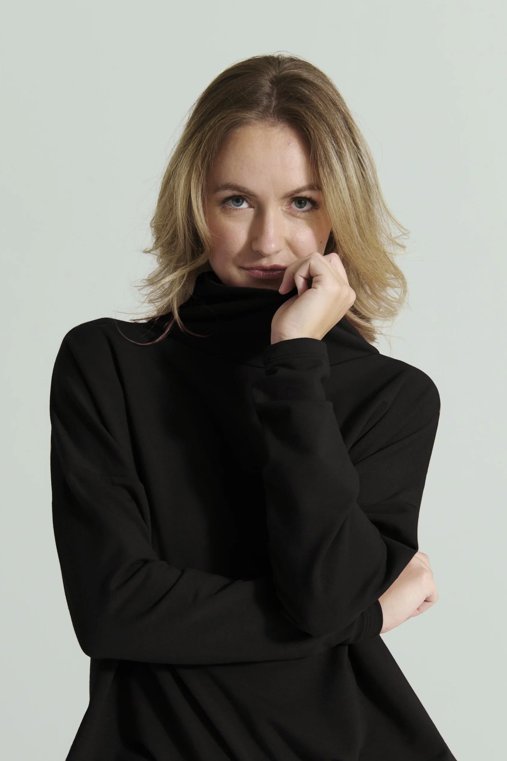 Waist up view of a woman wearing the Romaine Sweater by Message Factory in Black, a bamboo/organic cotton sweatshirt with a funnel neck , a loose fit, and long drop sleeves. She is standing in front of a white background. 