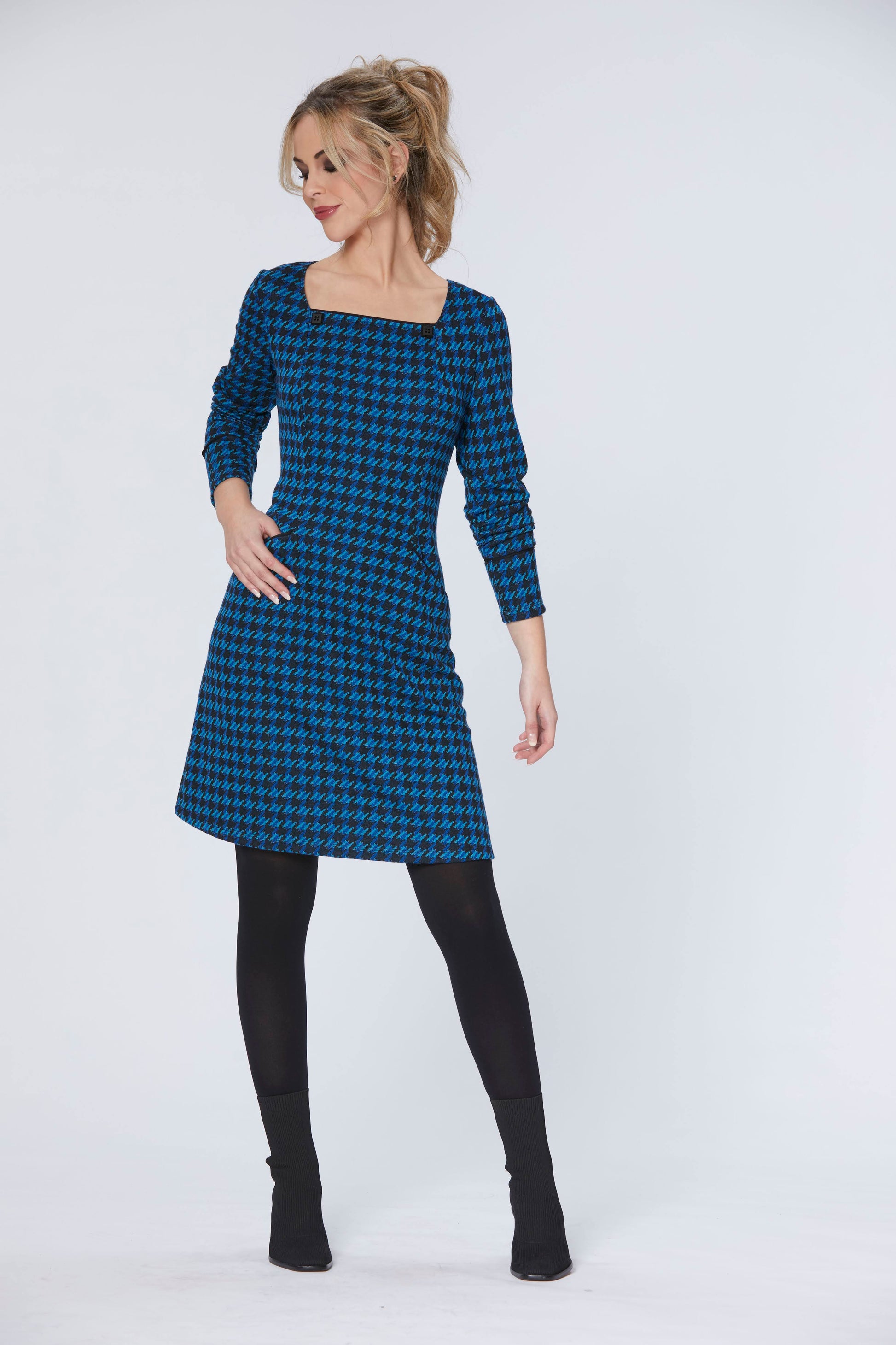 A woman wearing the Charing Dress by Luc Fontaine in Blue Houndstooth, featuring a square neckline with button details, a slight A-line shape, front slit pockets, and an above-the-knee length. She's standing in front of a white background. 