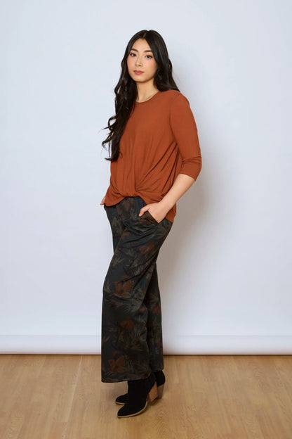 A woman wearing the Luisa Pants by Tangente in Floral, featuring an elastic waist with pleats, straight legs, and side pockets. She is wearing them with a rust coloured top and standing in front of a white background. 