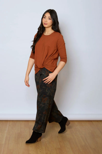 A woman wearing the Luisa Pants by Tangente in Floral, featuring an elastic waist with pleats, straight legs, and side pockets. She is wearing them with a rust coloured top and standing in front of a white background. 