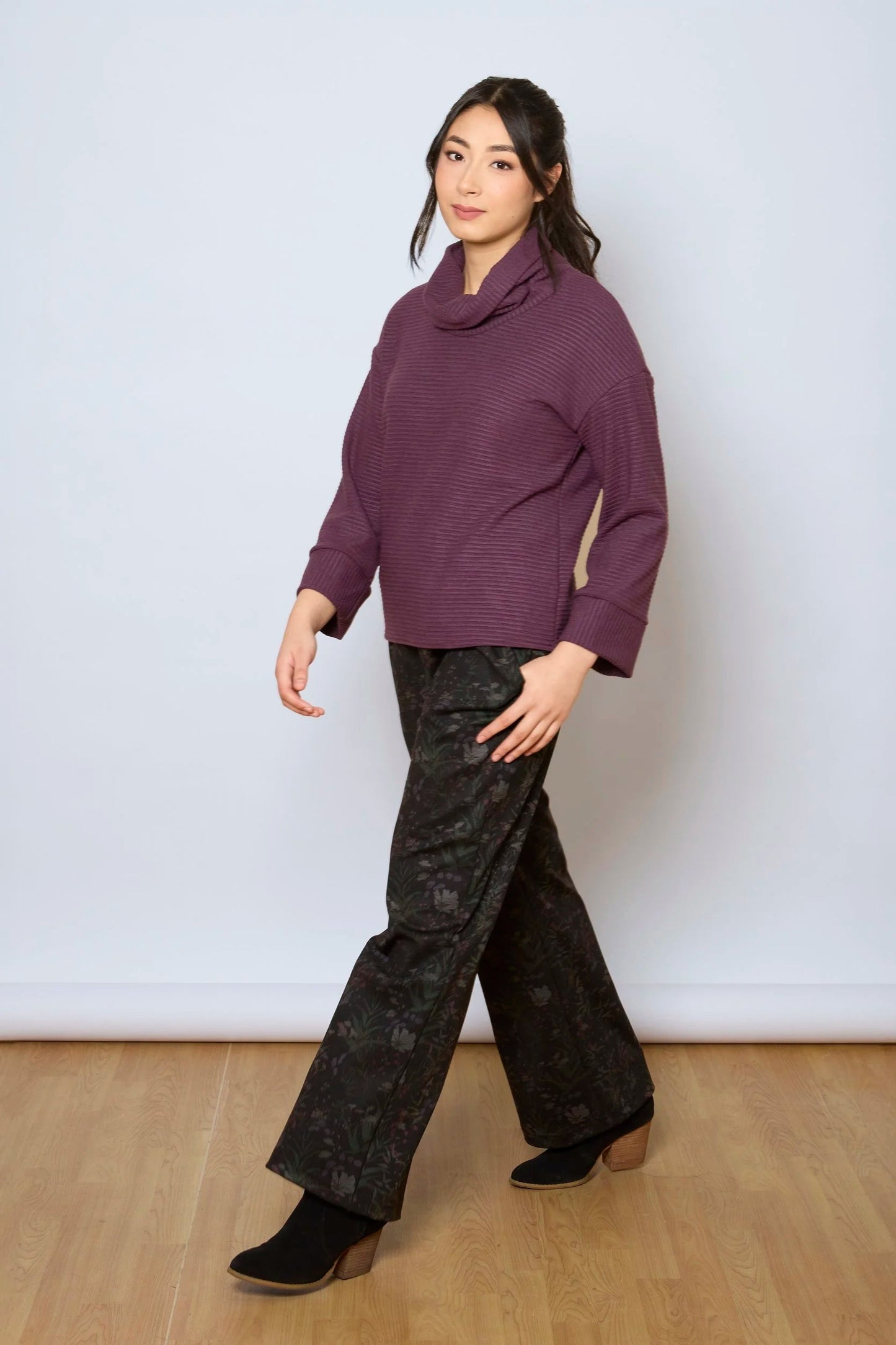A woman wearing the Luisa Pants by Tangente in Woodland print, featuring an elastic waist with pleats, straight legs, and side pockets. She is wearing them with a purple top and standing in front of a white background. 