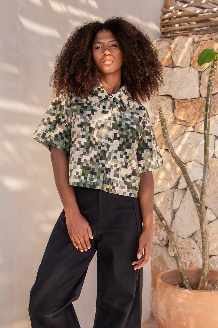 A woman wearing the Almeria Top by MAS in Camo, a slightly cropped button up short sleeved shirt with a large collar and a pixellated print in green and earth tones. She's wearing it with the Vienna Pants in Black, and standing outside by a stone wall. 
