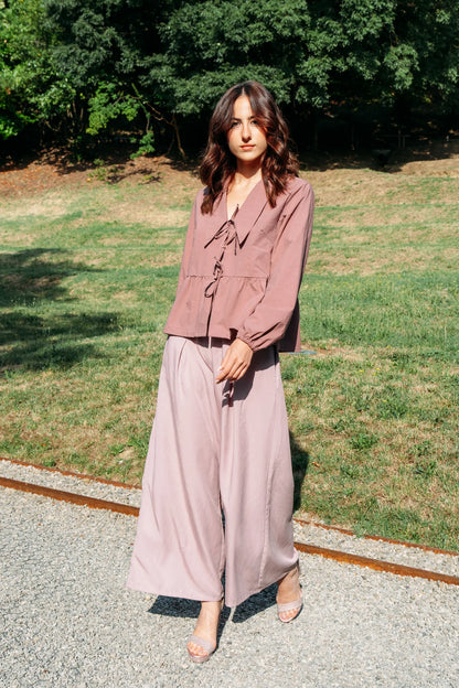 A woman wearing the Siena Pants by MAS in Raspberry, wide legged trousers with an elastic waist. She is wearing them with a pink jacket and standing outdoors. 