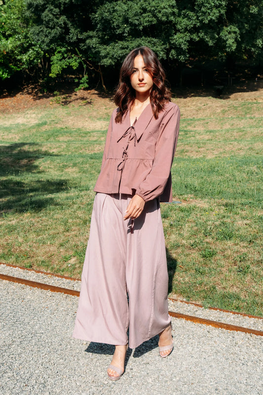A woman wearing the Siena Pants by MAS in Raspberry, wide legged trousers with an elastic waist. She is wearing them with a pink jacket and standing outdoors. 