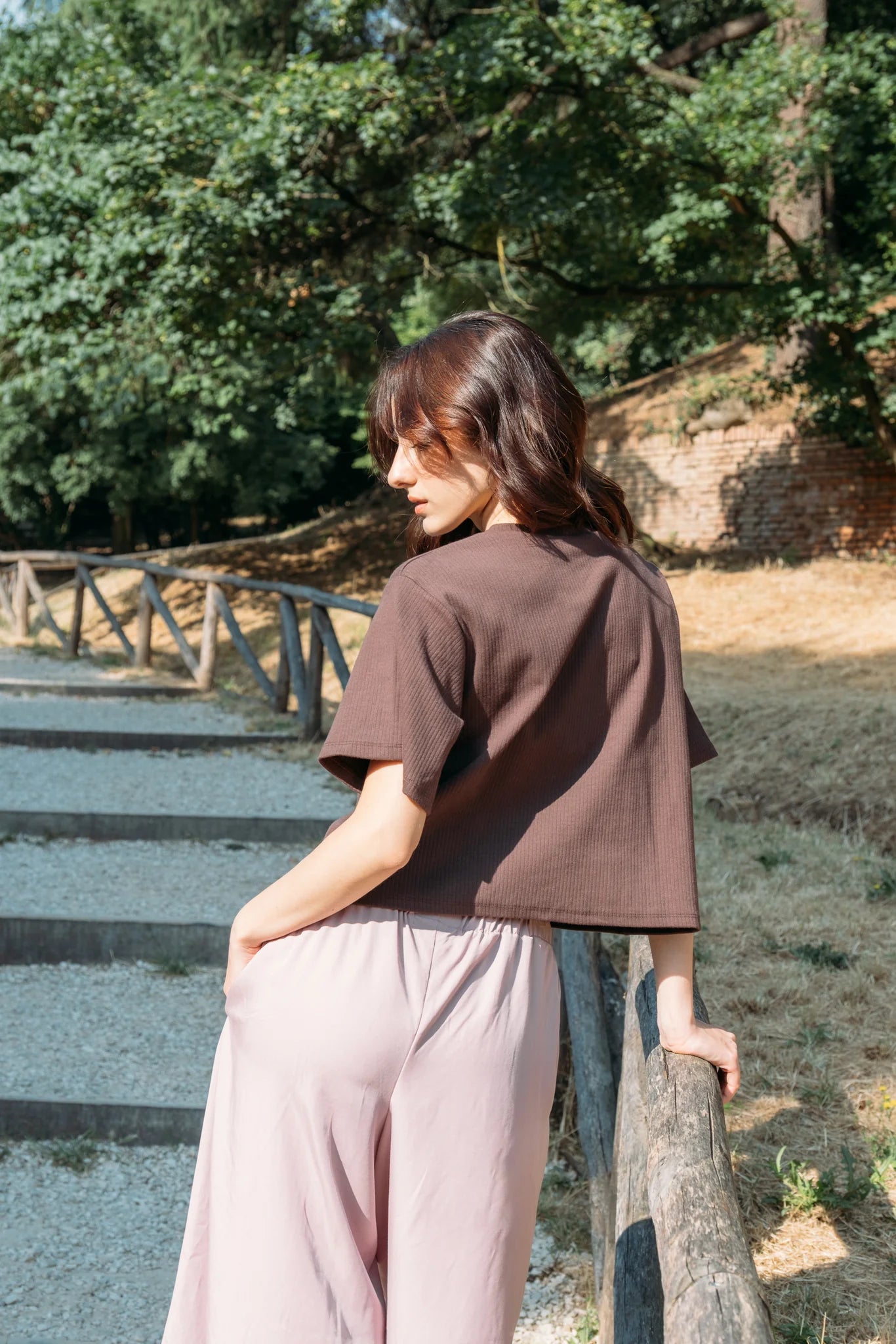Back view of a woman wearing the Siena Pants by MAS in Raspberry, wide legged trousers with an elastic waist. She is wearing them with a brown top and standing outdoors on a fenced pathway.
