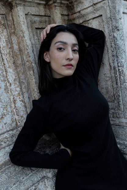 Woman in wearing the Mecca Dress by MAS in Black, a long slim-fitting  turtleneck dress. She is standing next to a stone wall.