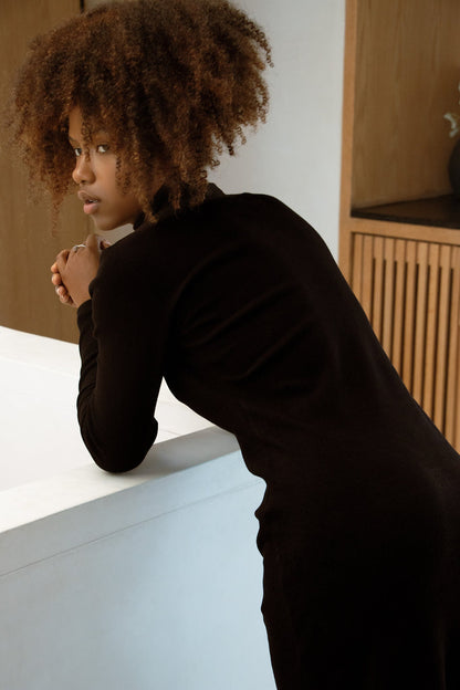 Back view of a woman in wearing the Mecca Dress by MAS in Pepper, a long slim-fitting  turtleneck dress. She is leaning over a white countertop.