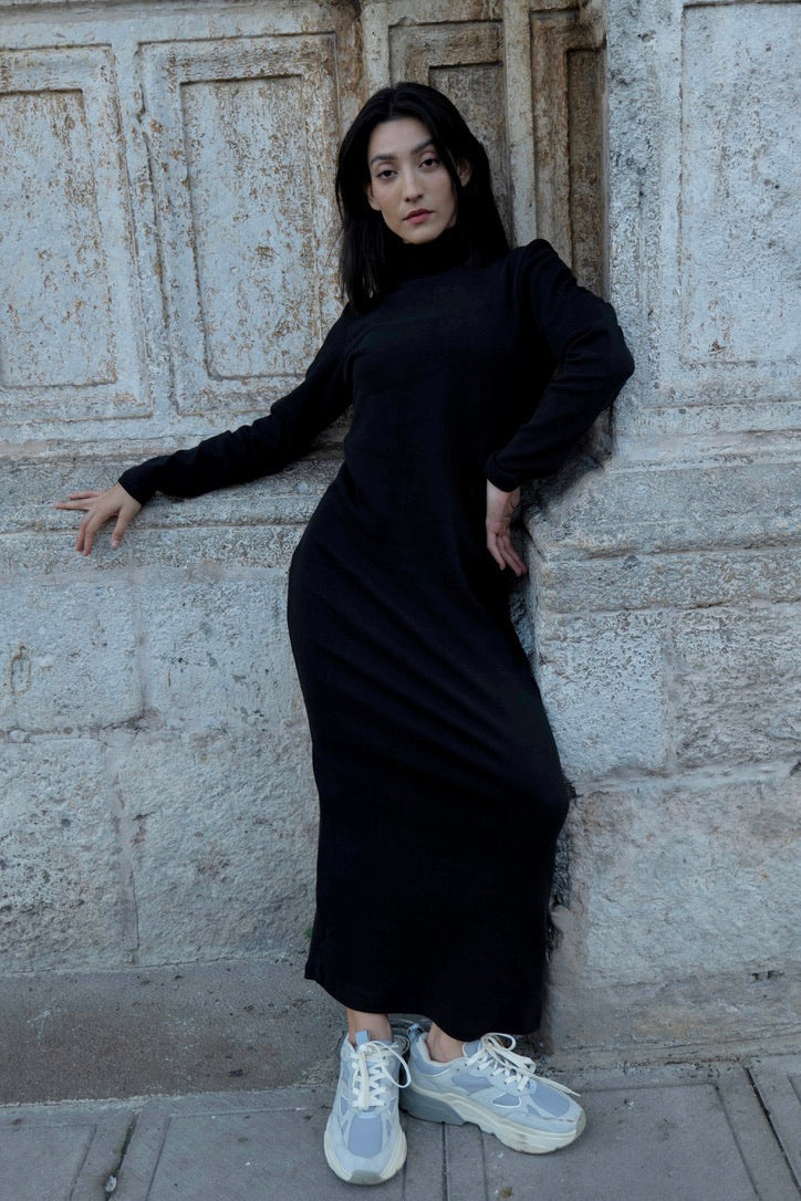 Woman in wearing the Mecca Dress by MAS in Pepper, a long slim-fitting  turtleneck dress. She is standing next to a stone wall.