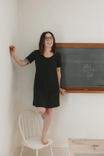 A woman wearing the Meadow Dress in Slub by Blondie Apparel in Black, standing on a white chair in front of a chalkboard