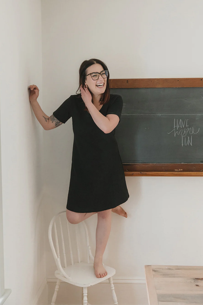 A woman wearing the Meadow Dress in Slub by Blondie Apparel in Black, standing on a white chair in front of a chalkboard