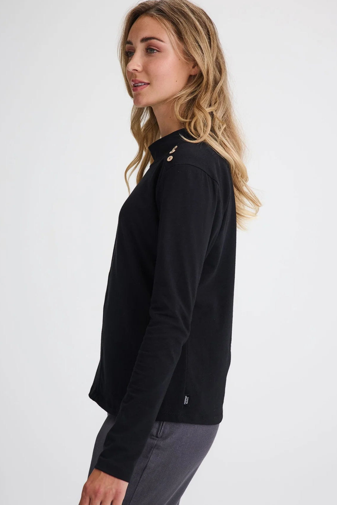 Side view of a woman wearing the Carleton Top by Message Factory in a Black, a long-sleeve top with button details on the shoulder. She is wearing it with grey bottoms and standing in front of a white background. 