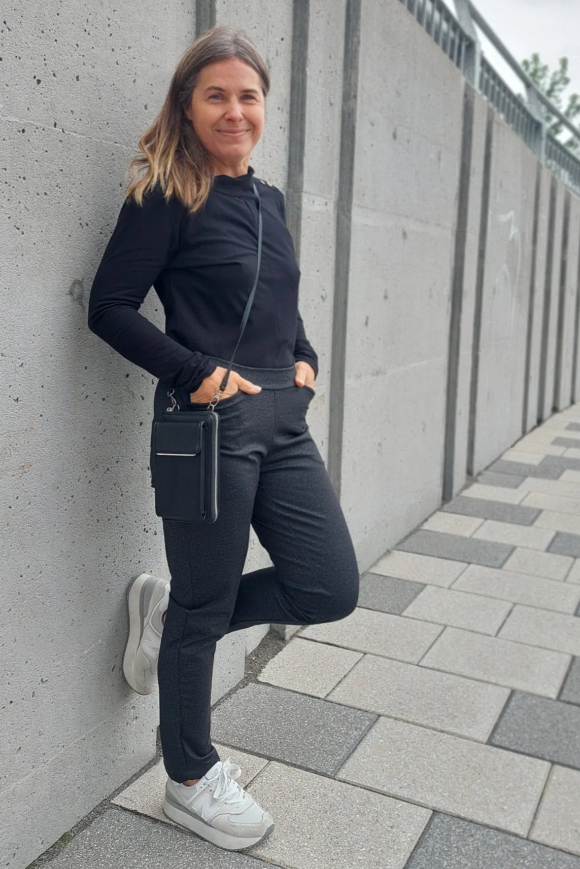 A woman wearing the Carleton Top by Message Factory in a Black, a long-sleeve top with button details on the shoulder. She is wearing it with black pants and standing outside leaning against a cement wall. 