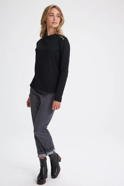 A woman wearing the Carleton Top by Message Factory in a Black, a long-sleeve top with button details on the shoulder. She is wearing it with grey bottoms and standing in front of a white background. 