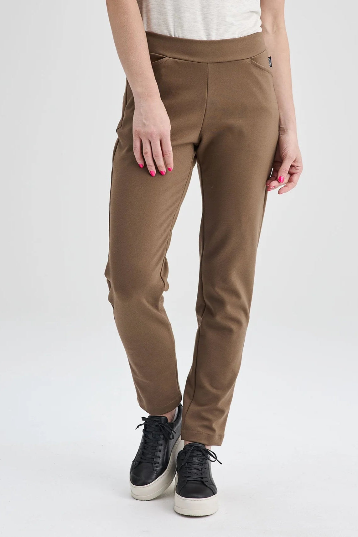 Waist down view of a woman wearing the Sherbrooke Pants by Message Factory in Caramel, pull-on stretch pants with a slim fit, smooth waistband, and two pockets. She is wearing them with a white top and standing in front of a white background. 