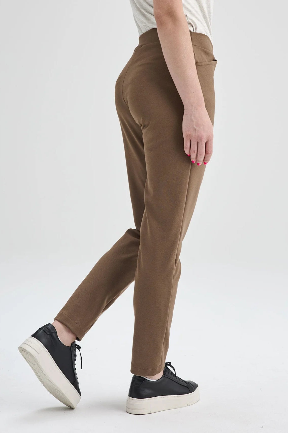 Side waist down view of Waist down view of a woman wearing the Sherbrooke Pants by Message Factory in Caramel, pull-on stretch pants with a slim fit, smooth waistband, and two pockets. She is wearing them with a white top and standing in front of a white background. 