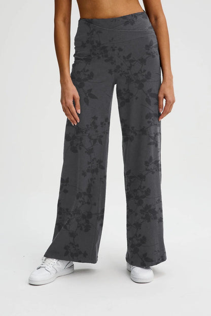 Waist down view of a woman wearing the Villeray Palazzo Pants by Message Factory in Grey Floral Print, wide legged organic cotton pants with a pull-on elastic waistband with a cross-over detail. She is standing in front of a white background. 