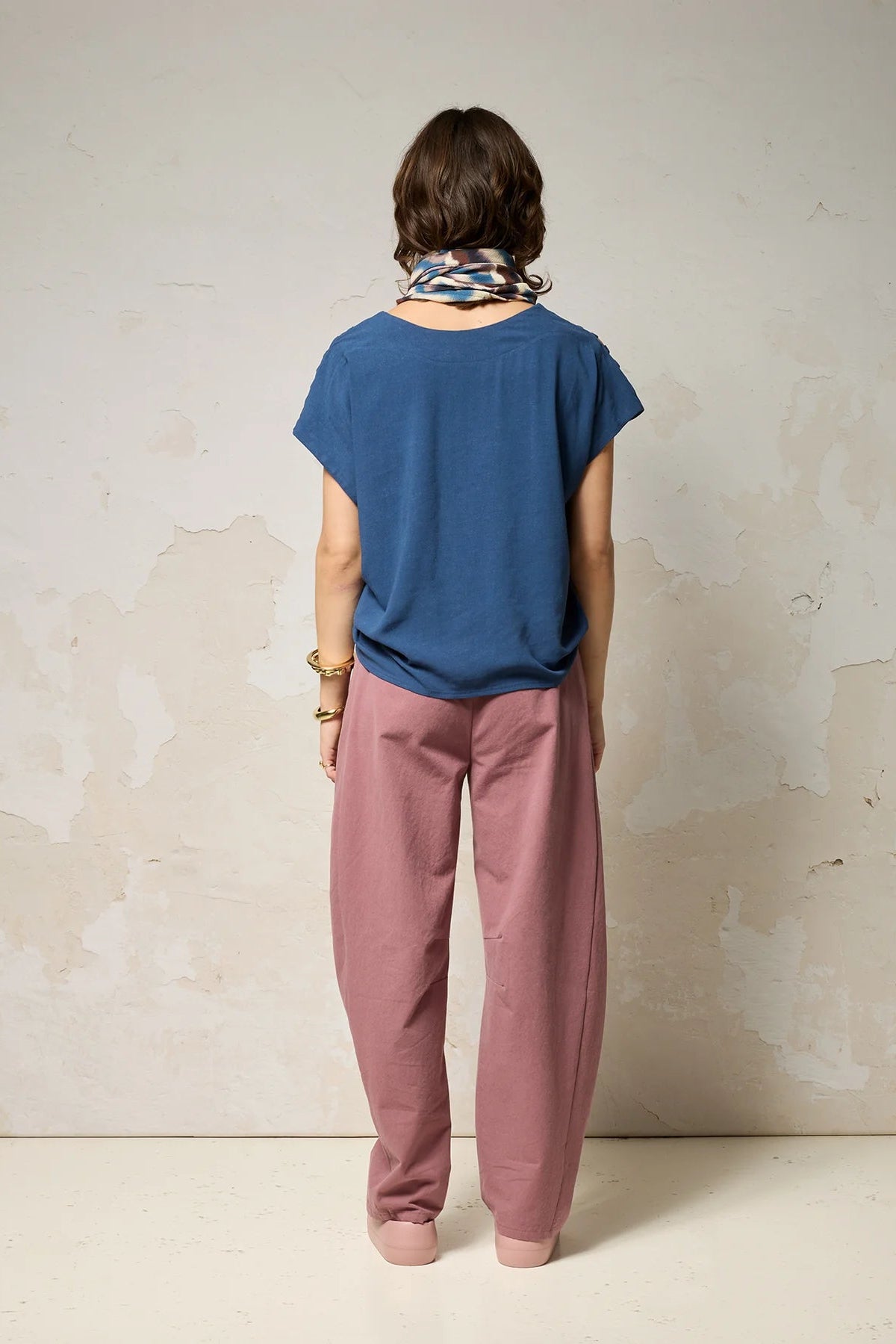 Back view of a woman wearing the Mia Top by Cokluch in Dahlia pink, a short sleeved top with openwork ladder-like cutouts at the shoulders, a loose fit, and V-neck. She is wearing it with black pants and standing against a plain background. 