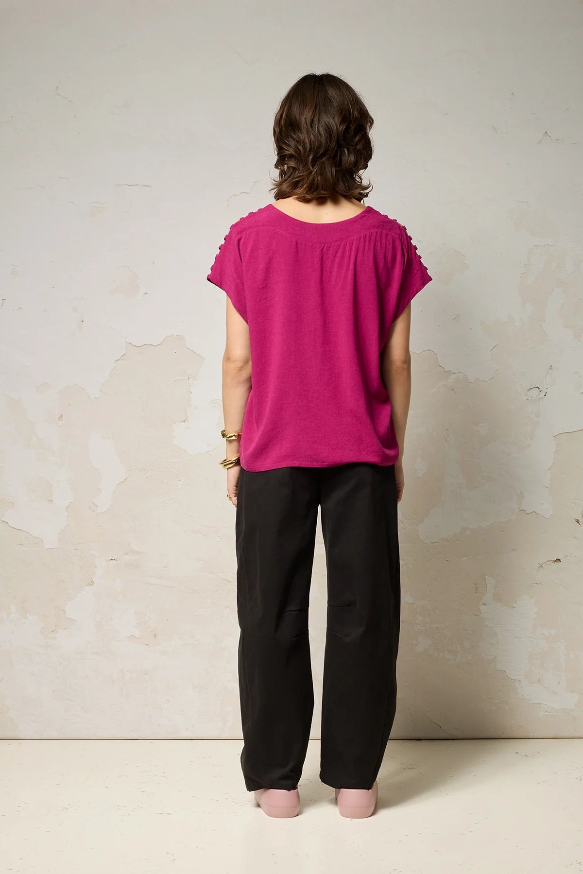 Back view of a woman wearing the Mia Top by Cokluch in Dahlia pink, a short sleeved top with openwork ladder-like cutouts at the shoulders, a loose fit, and V-neck. She is wearing it with black pants and standing against a plain background. 