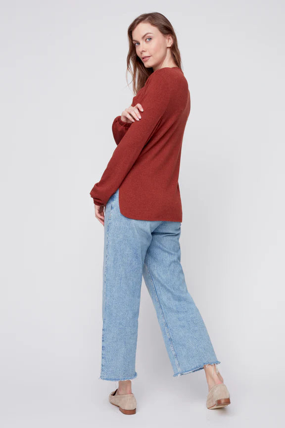 Back view of a woman wearing the Monica Sweater by Julei in Ochre, featuring a modal knit, a twist detail at the neck, bloused sleeves, and side slits. She is wearing it with jeans and standing in front of a white background. 