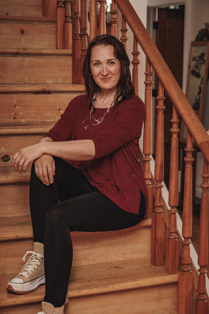 Woman wearing the Emmy Top by Moovment in Burgundy, with a boat neck, 3/4 sleeves, and side slits. She is wearing it with black pants and sitting on a wooden staircase.