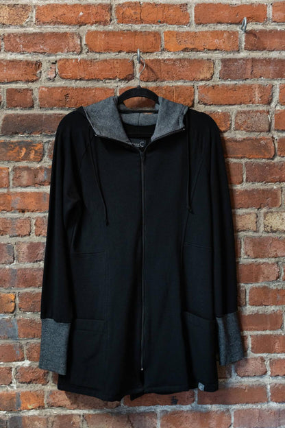The Calma Jacket by Marie C in Black, a mid-thigh length jacket with a hood and contrasting cuffs and lining, is shown hanging against a brick wall. 