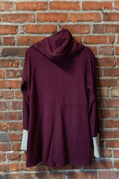 The Calma Jacket by Marie C in Wine, a mid-thigh length jacket with a hood and contrasting cuffs and lining, is shown hanging against a brick wall. 