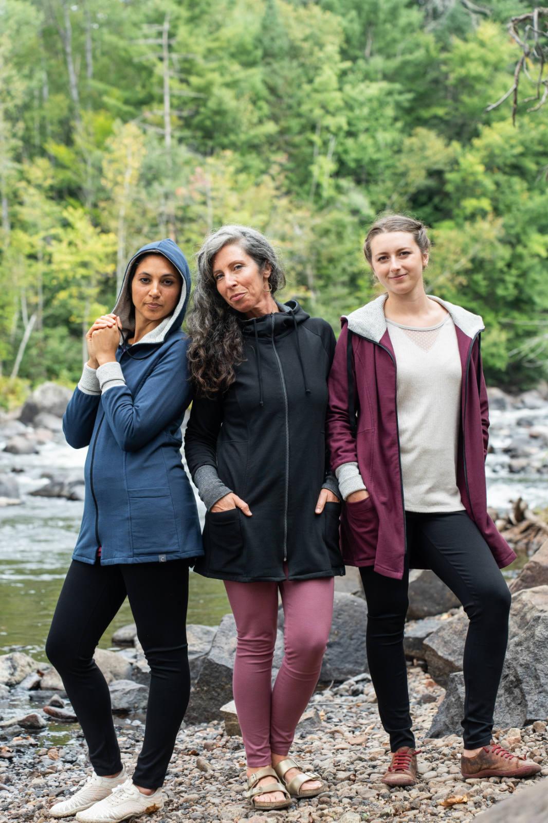 Three women wearing the Calma Jacket by Marie C in Navy, Black and Wine, a mid-thigh length jacket with a hood and contrasting cuffs and lining. They are standing outside by a river. 