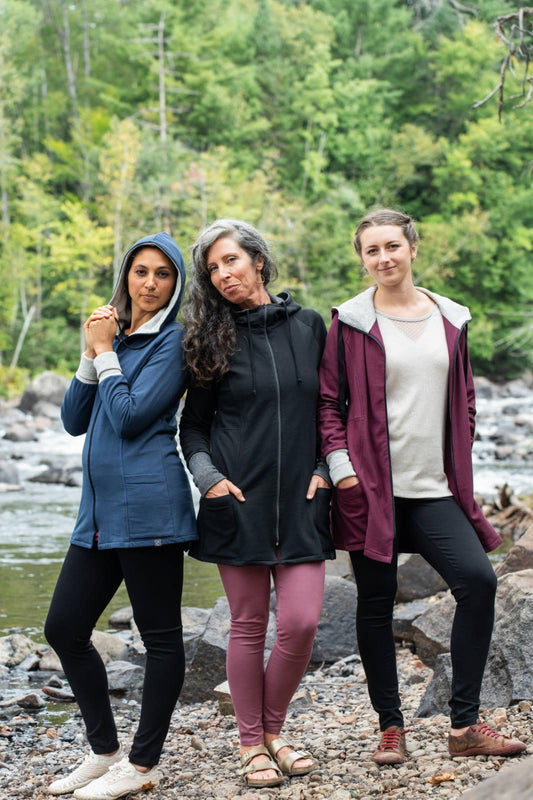 Three women wearing the Calma Jacket by Marie C in Navy, Black and Wine, a mid-thigh length jacket with a hood and contrasting cuffs and lining. They are standing outside by a river. 