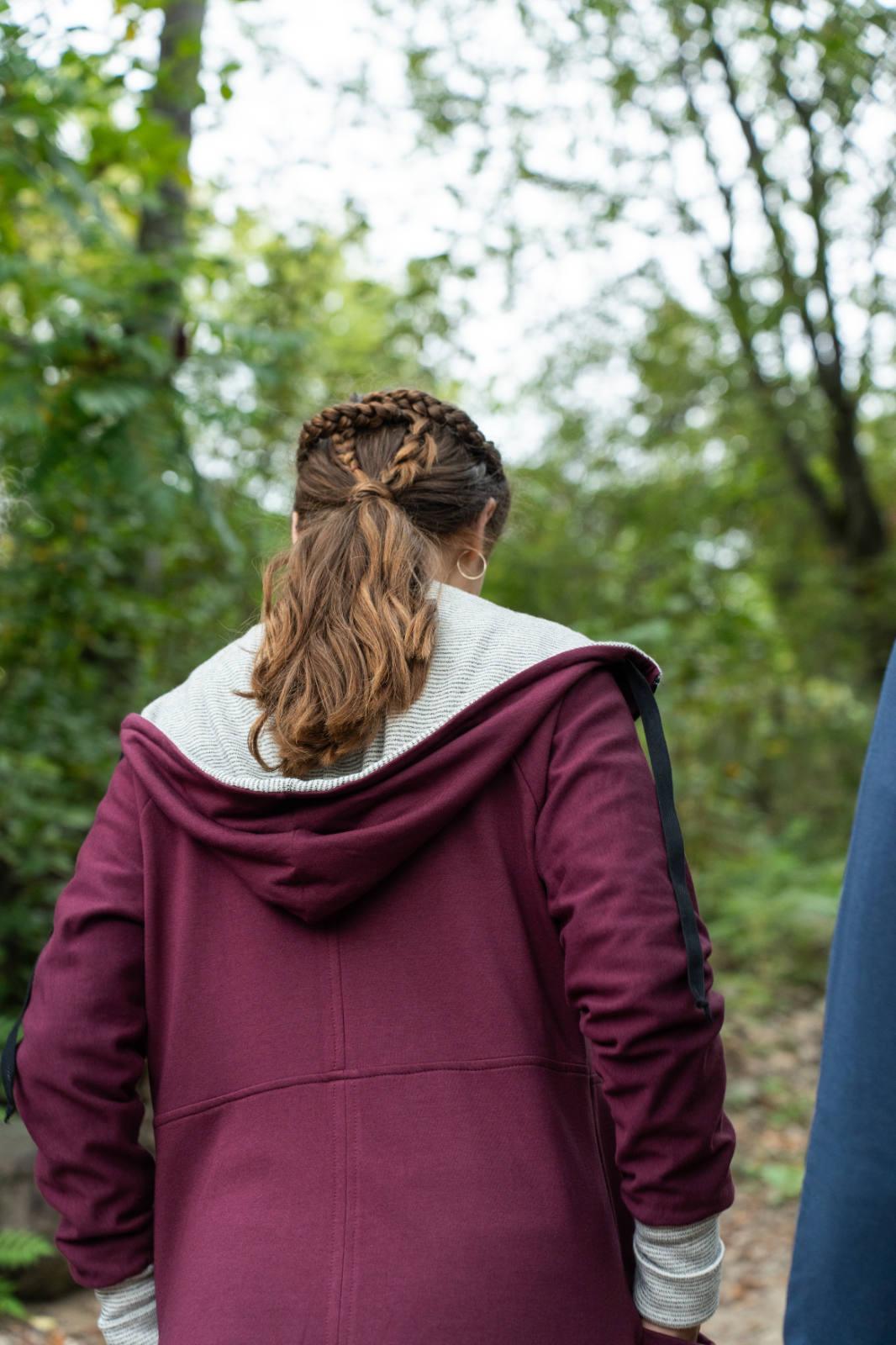 Back view of a woman wearing the Calma Jacket by Marie C in Wine, a mid-thigh length jacket with a hood and contrasting cuffs and lining. She is walking in a forest. 