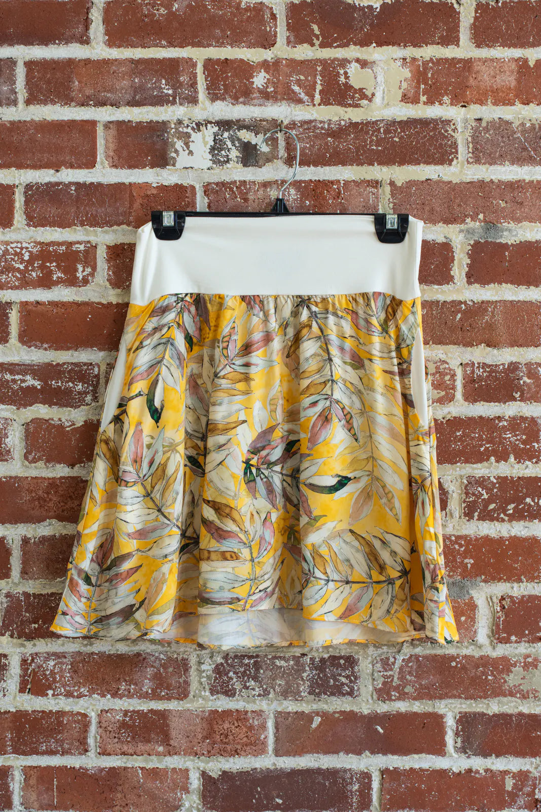 The Soleil Skirt by Marie C in Yellow Floral, a short A-line skirt with a contrasting white pull on waistband, is shown hanging from a brick wall. 