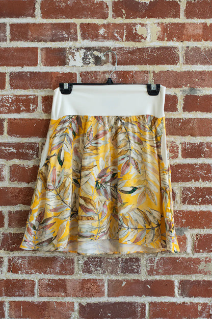 The Soleil Skirt by Marie C in Yellow Floral, a short A-line skirt with a contrasting white pull on waistband, is shown hanging from a brick wall. 