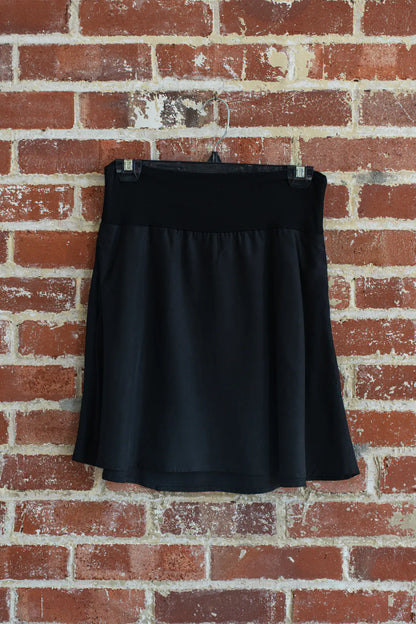 The Soleil Skirt by Marie C in Black Floral, a short A-line skirt with a pull on waistband, is shown hanging from a brick wall. 